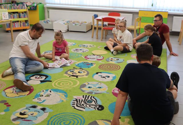 Na zdjęciu znajduje się trójka mężczyzn oraz piątka dzieci i słuchają książki, którą czyta mężczyzna po lewej. W tle znajdują się półki z książkami, stoliki z krzesłami.