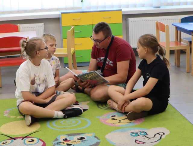 Na zdjęciu znajduje się mężczyzna z trójką dzieci na dywanie i czyta książkę, w tle znajdują się stoliki, szafka.