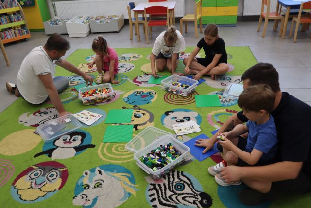 Na zdjęciu znajduje się trójka mężczyzn i piątka dzieci na dywanie i układają klocki LEGO, które znajdują się w plastikowych pudełkach.