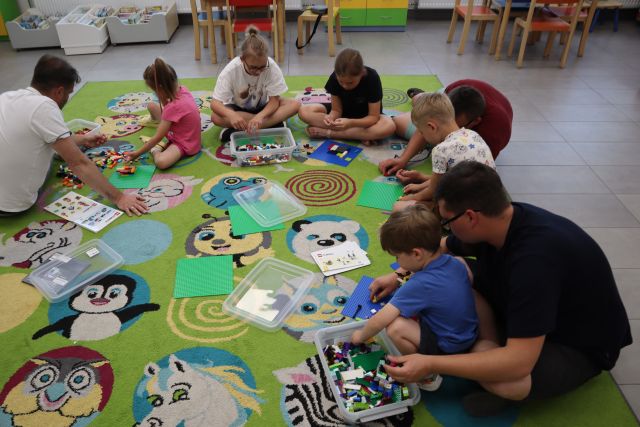Na zdjęciu znajduje się piątka dzieci i dwóch mężczyzn i układają klocki LEGO na dywanie. W tle znajdują się stoliki, przesuwane półki z książkami.