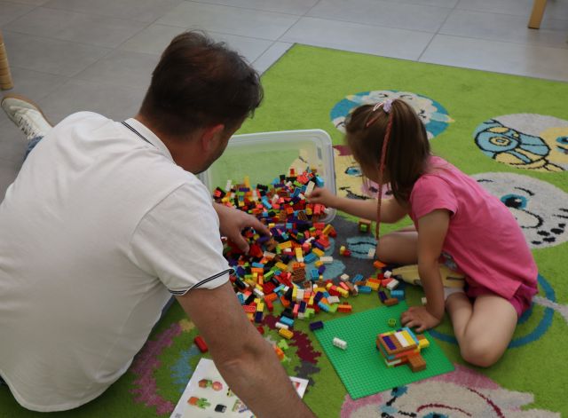 Na zdjęciu znajduje się mężczyzna wraz z dziewczynką i wyciągają klocki z przechylonego plastikowego pojemnika. Klocki układają na dywanie, a zdjęcie zostało wykonane od tyłu.