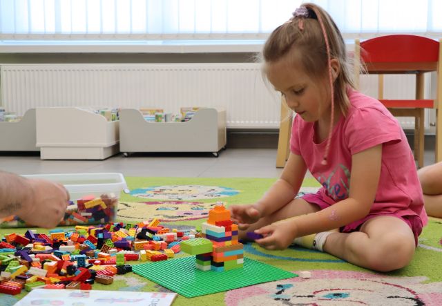 Na zdjęciu po prawej stronie znajduje się dziewczynka siedząca na dywanie po lewej stronie są rozłożone klocki LEGO, z których dziewczynka buduje, uchwycona na zdjęciu została również dłoń mężczyzny. W tle znajdują się jeżdżące półki z książkami, okna oraz krzesełka.