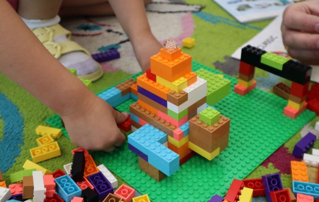 Na zdjęciu znajduje się konstrukcja zbudowana z klocków LEGO na zielonej podkładce, jest od lewej strony trzymana przez dziewczynkę, której uchwycone zostały tylko ręce. Praca leży na dywanie.