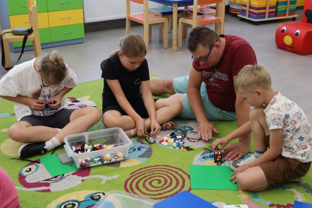 Na zdjęciu znajduje się mężczyzna wraz z trójką dzieci i wspólnie budują klocki LEGO na dywanie, w tle znajdują się krzesełka, pufa.