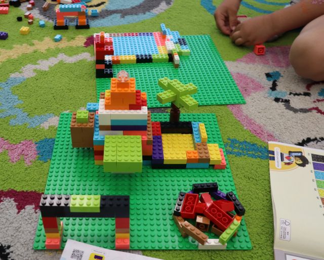 Na zdjęciu znajduje się zbudowany z klocków LEGO na zielonych podkładkach budynek, ognisko, drzewa, basen. Po prawej stronie leży instrukcja oraz znajdują się dłonie dziewczynki, która buduje kolejne elementy.