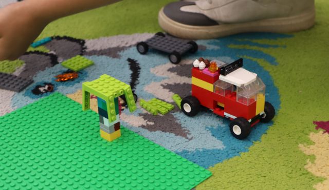 Na zdjęciu znajduje się leżąca na podłodze podkładka LEGO, na której zbudowane jest drzewo, obok leży zbudowany czerwony pojazd oraz but chłopca.