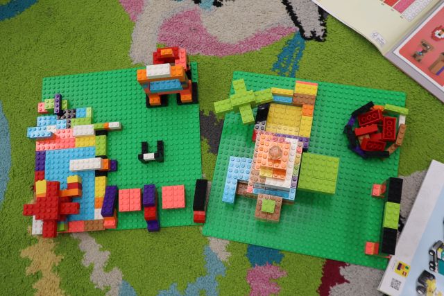 Na zdjęciu wykonanym od góry znajdują się dwie zielone podkładki LEGO na dywanie, na których zbudowany jest budynek, drzewa, robot, basen, ognisko, po prawej stronie znajdują się dwie instrukcje.