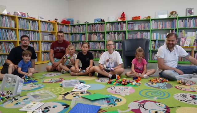 Na zdjęciu znajduje się trójka mężczyzn oraz piątka dzieci i siedzą na dywanie do wspólnego zdjęcia wraz ze swoimi konstrukcjami z LEGO, na dywanie przed nimi leżą podkładki, klocki, a w tle znajdują się półki z książkami, fotel oraz wystawka książek na stoliku.