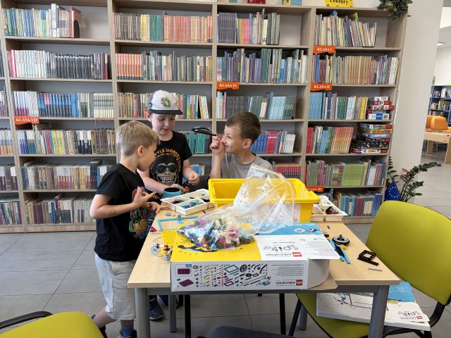 Na zdjęciu znajduje się trójka chłopców przy stoliku, na którym rozłożone są klocki LEGO, karton. Jeden chłopiec siedzi i pokazuje stojącemu obok koledze swoją konstrukcje z klocków. W tle znajdują się półki z książkami.