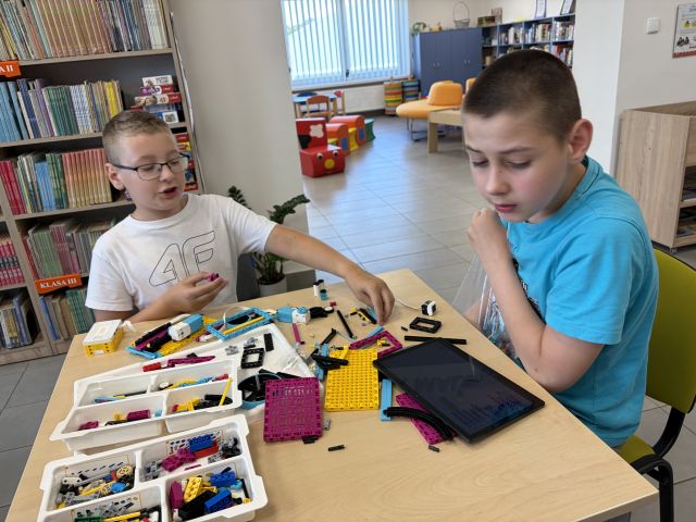 Na zdjęciu znajduje się dwójka chłopców, którzy układają klocki LEGO przy stoliku, przed chłopcem po prawo leży tablet. W tle znajdują się półki z książkami oraz wejście do drugiego pomieszczenia gdzie znajdują się pufy.