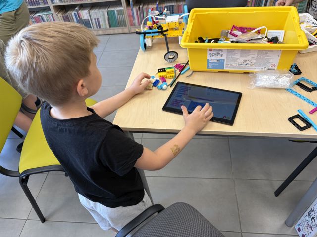 Na zdjęciu znajduje się stojący przy stoliku chłopiec, przed nim leży tablet na którym koduje zbudowane wcześniej auto, na stoliku leżą również klocki. Przy stoliku stoi zielone krzesło. Na przeciwko znajdują się półki z książkami.