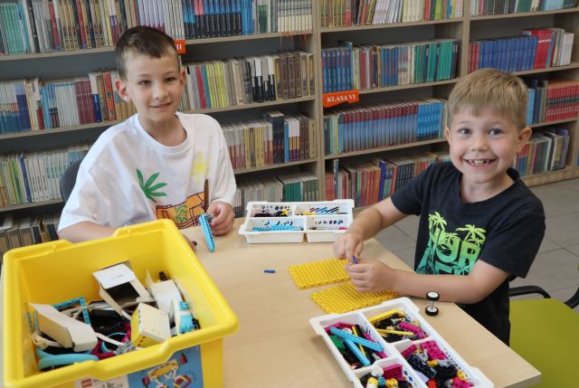 Na zdjęciu znajduje się dwójka chłopców pru i wspólnie układają klocki LEGO, w tle znajdują się półki z książkami.