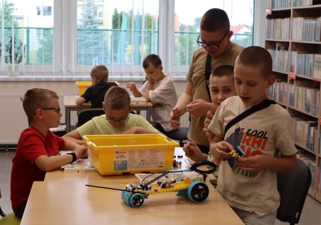 Na zdjęciu znajduje się siódemka chłopców przy stolikach i układają klocki LEGO, które znajdują się w pudełkach na stole. W tle znajdują się okna, a po prawej stronie półki z książkami.