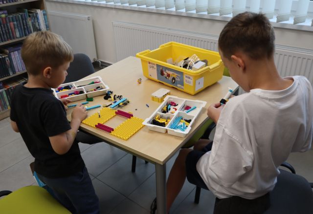 Na zdjęciu znajduje się dwójka chłopców, którzy układają wspólnie LEGO przy stoliku, w tle znajduje się ściana.