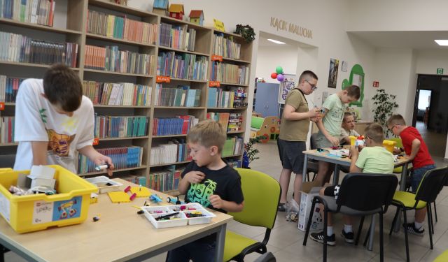 Na zdjęciu znajduje się pięciu chłopców przy stolikach i budują klocki LEGO, znajdujące się na stolikach. W tle znajdują się półki z książkami.
