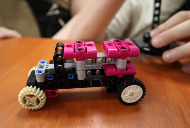 Na zdjęciu znajduje się mały samochód zbudowany z klocków LEGO, w tle znajduje się chłopiec.