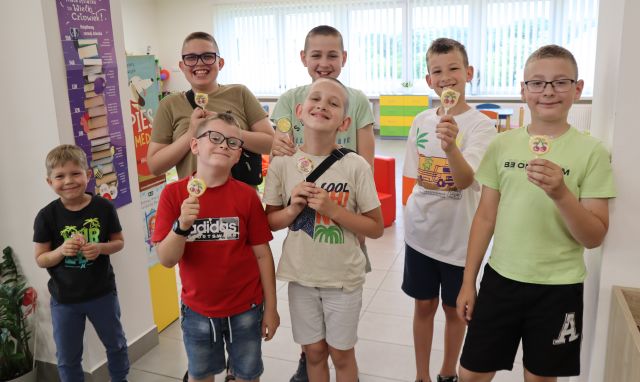 Na zdjęciu znajduje się siedmiu chłopców stojących do zdjęcia z lizakami, w tle znajdują się okna.