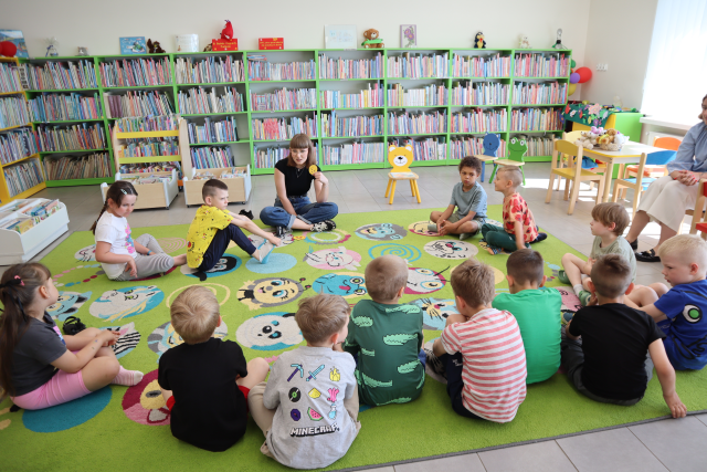 Na zdjęciu znajduje sie trzynaście dzieci siedzących na dywanie w okręgu na środku siedzi kobieta i pokazuje im papierowy emotikon żeby dzieci rozpoznały emocję. W tle znajdują się półki z książkami, małe krzesełka.