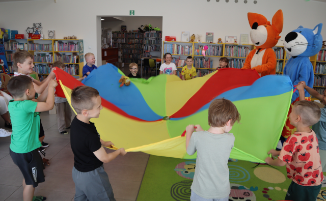 Na zdjęciu znajdują się dzieci wraz z maskotką lista i maskotką niebieskiego kota, wszyscy wspólnie bawią się chustą animacyjną, na której są maskotki. W tle znajdują się półki z książkami.
