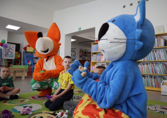 Na zdjęciu znajduje się niebieska maskotka kota, obok niej siedzą dzieci i pomarańczowa maskotka lisa, wszyscy trzymają po misiu. W tle znajdują się półki z książkami.