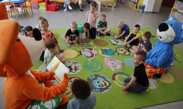 Na zdjęciu znajduje się pomarańczowa maskotka lisa, dookoła której na dywanie siedzą dzieci i niebieski kot. Maskotka lisa czyta dzieciom książkę. W tle znajdują się stoliki, pufy.