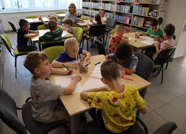 Na zdjęciu znajduje się trzynaście dzieci przy czterech stolikach i rysują na kartkach. Przy stoliku na końcu siedzi kobieta. W tle znajdują się półki z książkami i okna.