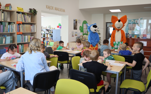 Na zdjęciu znajduje się trzynaście przedszkolaków i dwie kobiety przy czterech stołach i rysują na kartkach. W tle przed nimi stoi maskotka lisa oraz maskotka niebieskiego kota, po lewej stronie znajdują się półki z książkami.