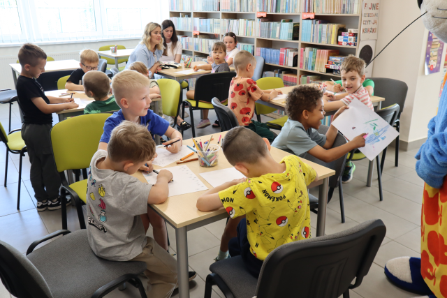 Na zdjęciu przy czterech stolikach siedzi trzynaście dzieci i rysują, przy jednym ze stolików siedzą również dwie kobiety. W tle znajdują się półki z książkami.