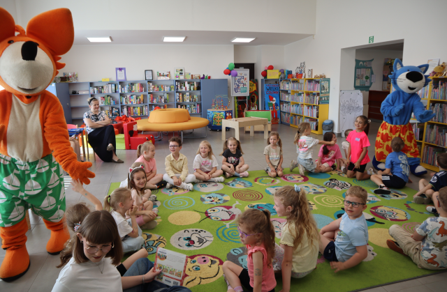 Na zdjęciu znajdują się dzieci na dywanie, dookoła dywanu chodzą dwie maskotki, po prawej niebieski kot, po lewej lis i przybijają piątki z przedszkolakami. W tle znajdują się półki z grami, okrągła sofa, stoliki.