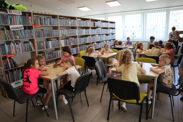 Na zdjęciu znajdują się przedszkolaki przy stolikach i kolorują, w tle znajdują się półki z książkami i okna.