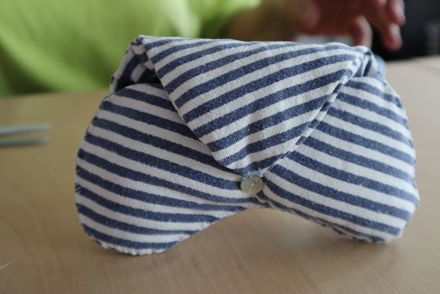 Na zdjęciu znajduje się etui na okulary uszyte z biało niebieskiego materiału w paski, praca leży na stole.