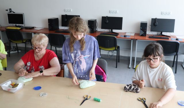 Na zdjęciu znajdują się dwie dziewczynki oraz kobieta przy złączonych stołach, dziewczynka na środku stoi . Uczestniczy zajęć szyją etui na okulary, na stolikach znajdują się nożyczki, nici. Warsztaty odbywają się w sali komputerowej.