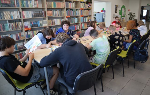 Na zdjęciu znajdują się uczniowie przy złączonych stołach i kolorują kwiatki dobrych manier, w tle znajdują się półki z książkami.