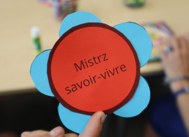 Na zdjęciu znajduje się kolorowy kwiat, który jest odznaką mistrza savoir vivre. W tle znajduje się stół oraz ręka uczestnika, który kończy swoją odznakę.