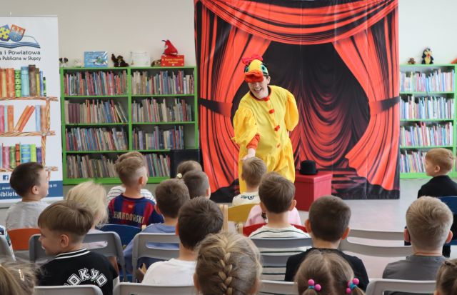 Na zdjęciu znajdują się dzieci na widowni, przed nimi stoi kobieta w stroju kaczki, w tle znajduje się czerwona kurtyna, półki z książkami oraz baner z logiem biblioteki i książkami po lewej stronie.