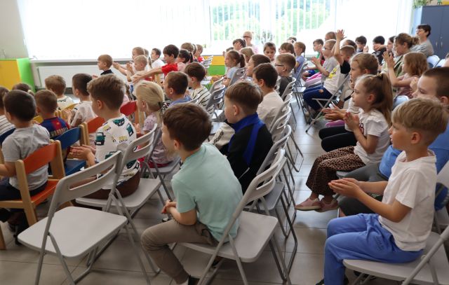 Na zdjęciu znajdują się dzieci siedzące na widowni w sali bibliotecznej i klaszczą, w tle znajdują się okna.