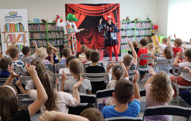 Na zdjęciu znajdują się dzieci na widowni, które powtarzają ruchy znajdującej się przed nimi przebranej kobiety z dużą igłą oraz mężczyzny przebranego za smoka. W tle znajduje się tablica z czerwoną kurtyną, baner z logiem biblioteki i książkami po lewej stronie oraz półki z książkami.