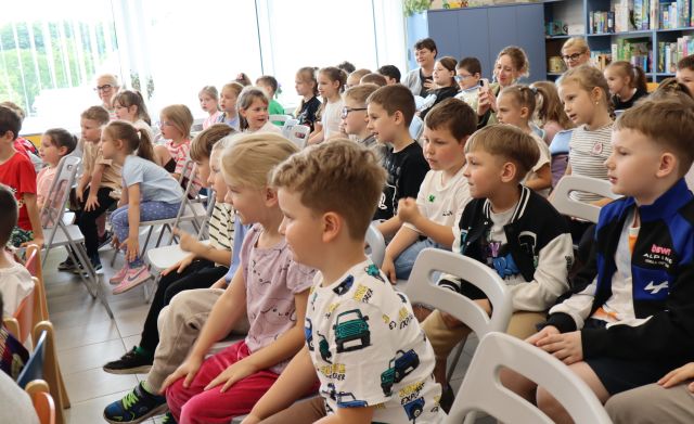 Na zdjęciu znajdują się dzieci na widowni w sali bibliotecznej, w tle znajdują się okna.