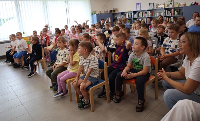 Na zdjęciu znajdują się dzieci na widowni w tle znajdują się okna, półki z grami i szafki.