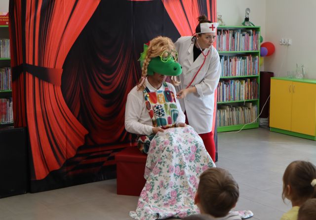 Na zdjęciu znajduje się siedzący na pufie mężczyzna przebrany za smoka, ma nogi przykryte kocem oraz perukę na głowie, obok niego stoi kobieta przebrana za lekarza i go bada. Za nimi znajduje się tablica z czerwoną kurtyną, w tle półki z książkami oraz szafka po prawej stronie.