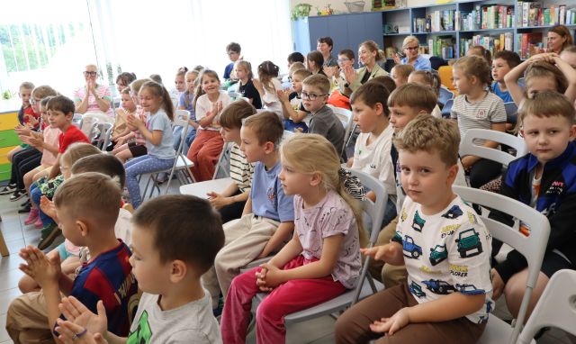 Na zdjęciu znajdują się dzieci siedzące na krzesłach na widowni, w tle znajdują się półki z grami oraz okna.