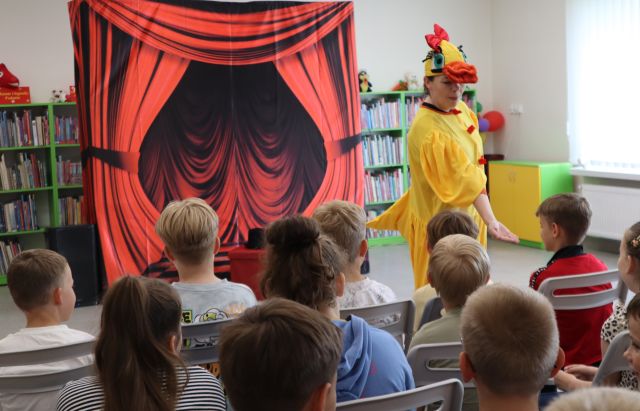 Na zdjęciu znajdują się uczniowie na widowni, a przed nimi stoi i wyciąga rękę do jednego ucznia kobieta w stroju kaczki. W tle znajduje się tablica z czerwoną kurtyną oraz półki z książkami.
