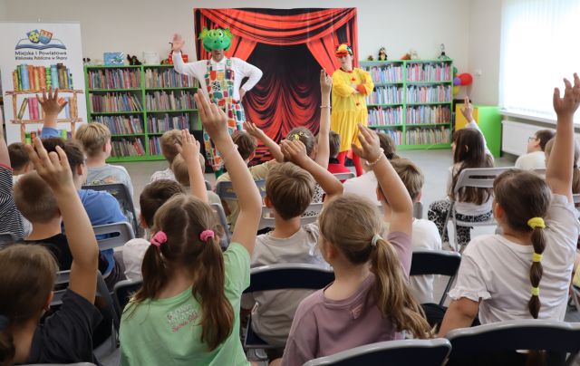 Na zdjęciu znajdują sie dzieci na widowni, mają podniesioną jedną rękę do góry, przed nimi stoi kobieta przebrana za kaczkę oraz mężczyzna przebrany za smoka, który również ma uniesioną jedną rękę do góry. W tle znajduje się tablica z czerwoną kurtyną oraz półki z książkami.