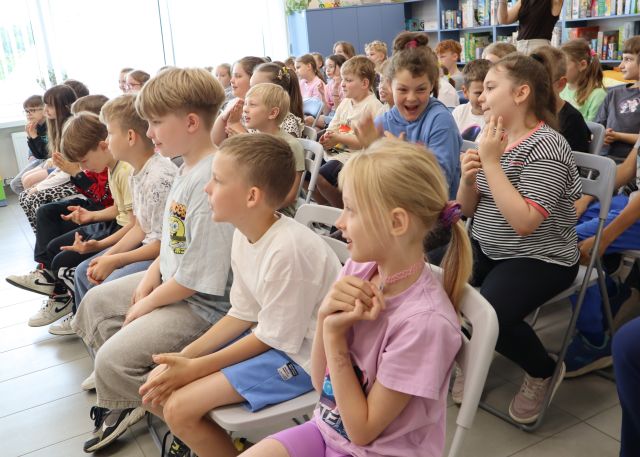 Na zdjęciu znajdują się uczniowie na krzesłach w sali bibliotecznej i oglądają spektakl. W tle znajdują się okna, po prawej stronie półki z grami, szafa.