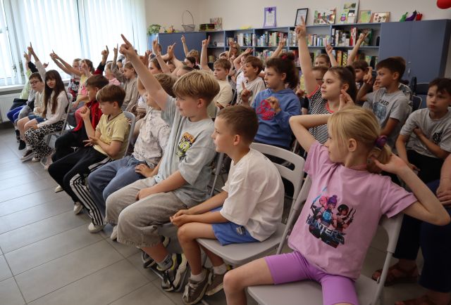 Na zdjęciu znajdują się uczniowie na krzesłach, część z nich ma uniesioną rękę do góry, w tle znajdują się okna, półki z grami, szafa.
