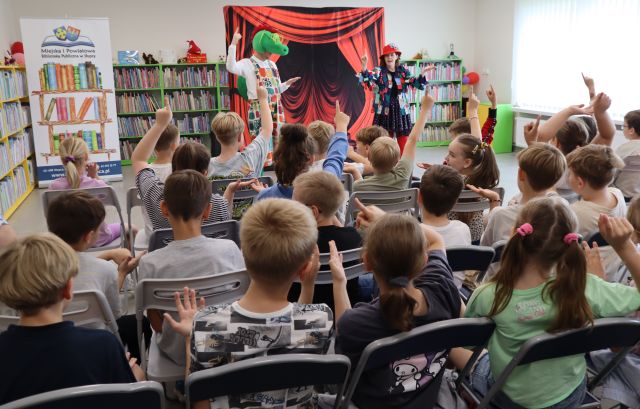 Na zdjęciu znajdują się uczniowie na krzesłach i powtarzają ruchy znajdujących się przed nimi aktorów, kobieta jest przebrana i ma w ręce dużą igłe, a stojący obok niej mężczyzna jest przebrany za smoka. W tle znajduje się tablica z czerwoną kotarą, baner z logiem biblioteki i książkami po lewej stronie oraz półki z książkami w tlr.