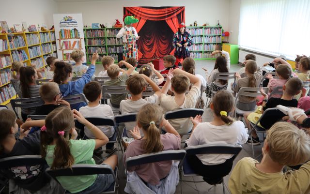 Na zdjęciu znajdują się uczniowie na krzesłach na widowni, częśc z nich ma delikatnie podniesioną rękę do góry, przed nimi stoi przebrana kobieta z dużą igłą oraz mężczyzna przebrany za smoka. W tle znajduje się tablica z czerwoną kurtyną, baner z książkami i logiem biblioteki po lewej stronie oraz półki z książkami.