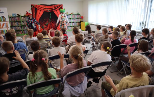 Na zdjęciu znajdują się uczniowie na krzesłach, przed nimi znajduje się przebrana kobieta, w jednej ręce trzyma dużą igłę, w drugiej długi sznurek oraz mężczyzna przebrany za smoka. W tle znajduje się tablica z czerwoną kurtyną, półki z książkami oraz okna po prawo.