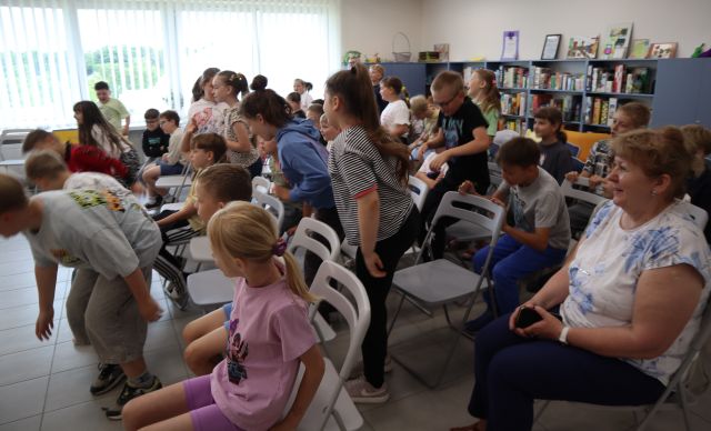 Na zdjęciu znajdują się dzieci na widowni, część z nich siedzi, część skacze. W tle znajdują się okna, półki z grami po prawej stronie.