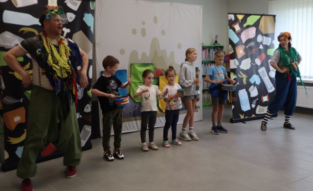 Na zdjęciu znajduje się piątka dzieci z instrumentami, po lewej stronie stoi mężczyzna przebrany za śmieciaka, a po prawej stronie kobieta. Za nimi znajdują się trzy tablice, dwie boczne są czarne z odpadkami, a środkowa z workami na śmieci.